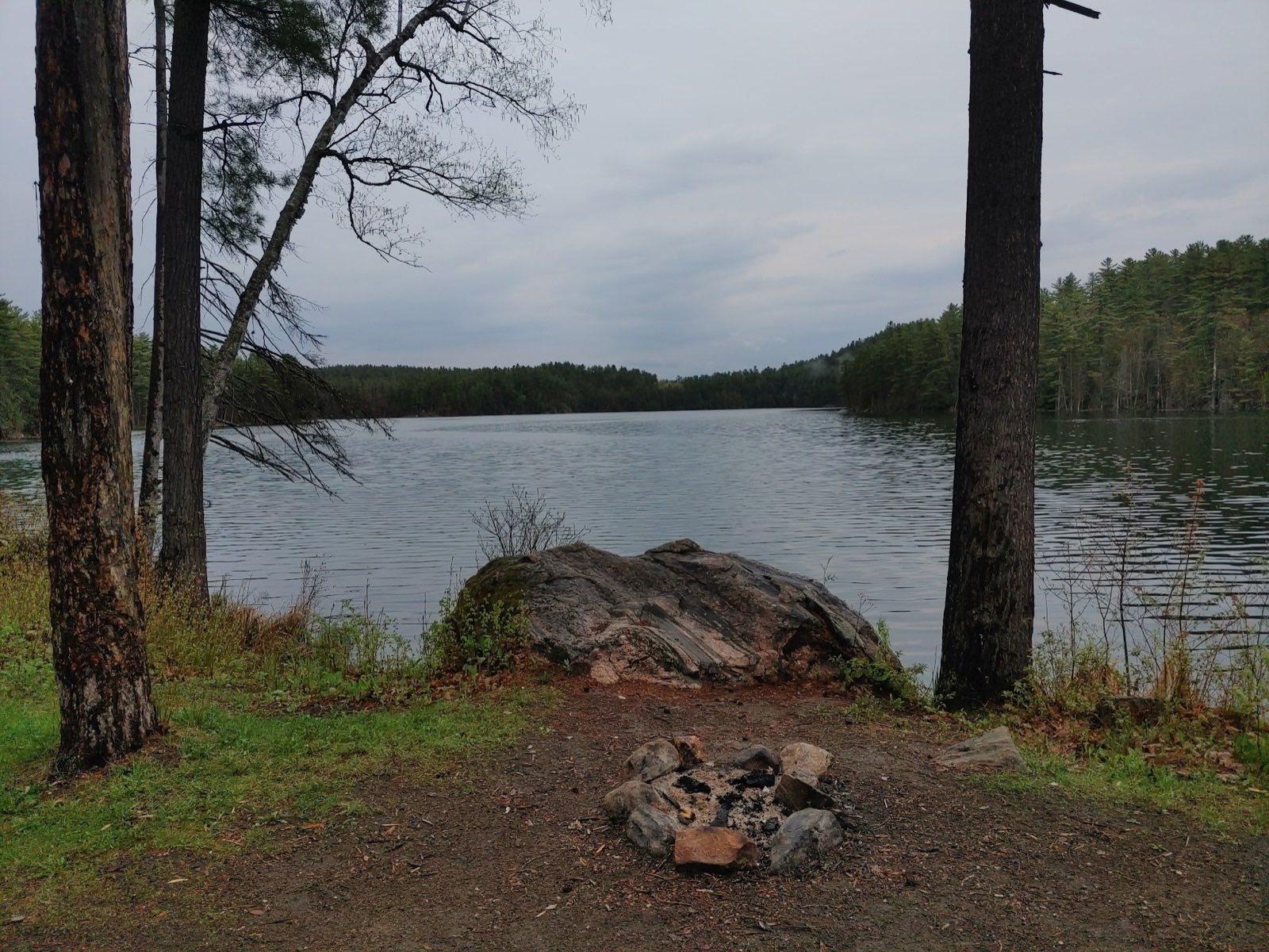 Crown Land campsite with a stone fire pit and boulder overlooking Wabun Lake on an overcast morning